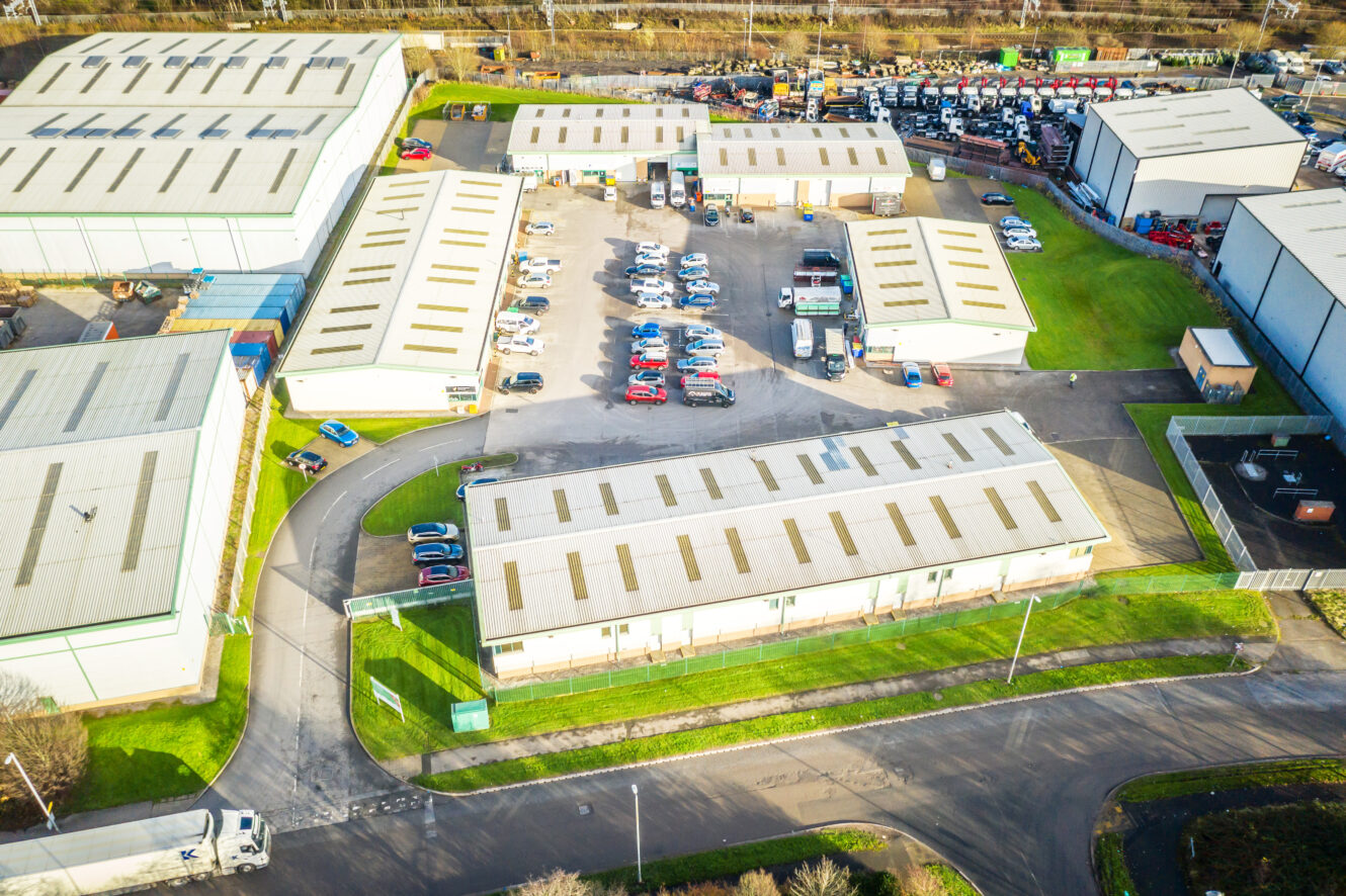 Aerial view of an industrial estate with multiple warehouse buildings, parked cars, and access roads surrounded by grassy areas.