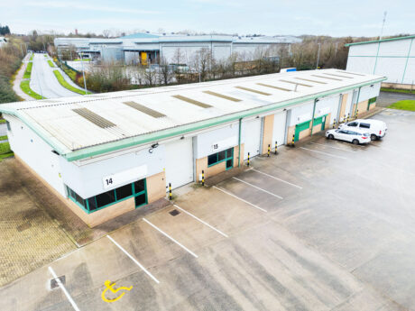A row of industrial warehouse units with numbered doors and parking spaces in front, including a designated accessible parking spot and two parked vehicles.