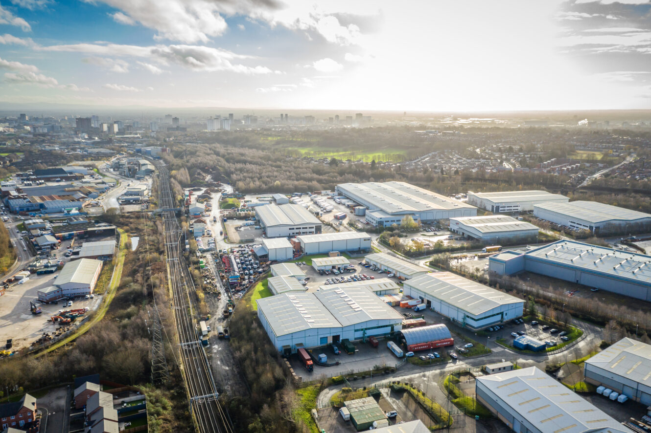 Aerial view of an industrial estate with warehouses, roads, and parked vehicles, adjacent to railroad tracks and surrounded by trees and urban areas under a partly cloudy sky.