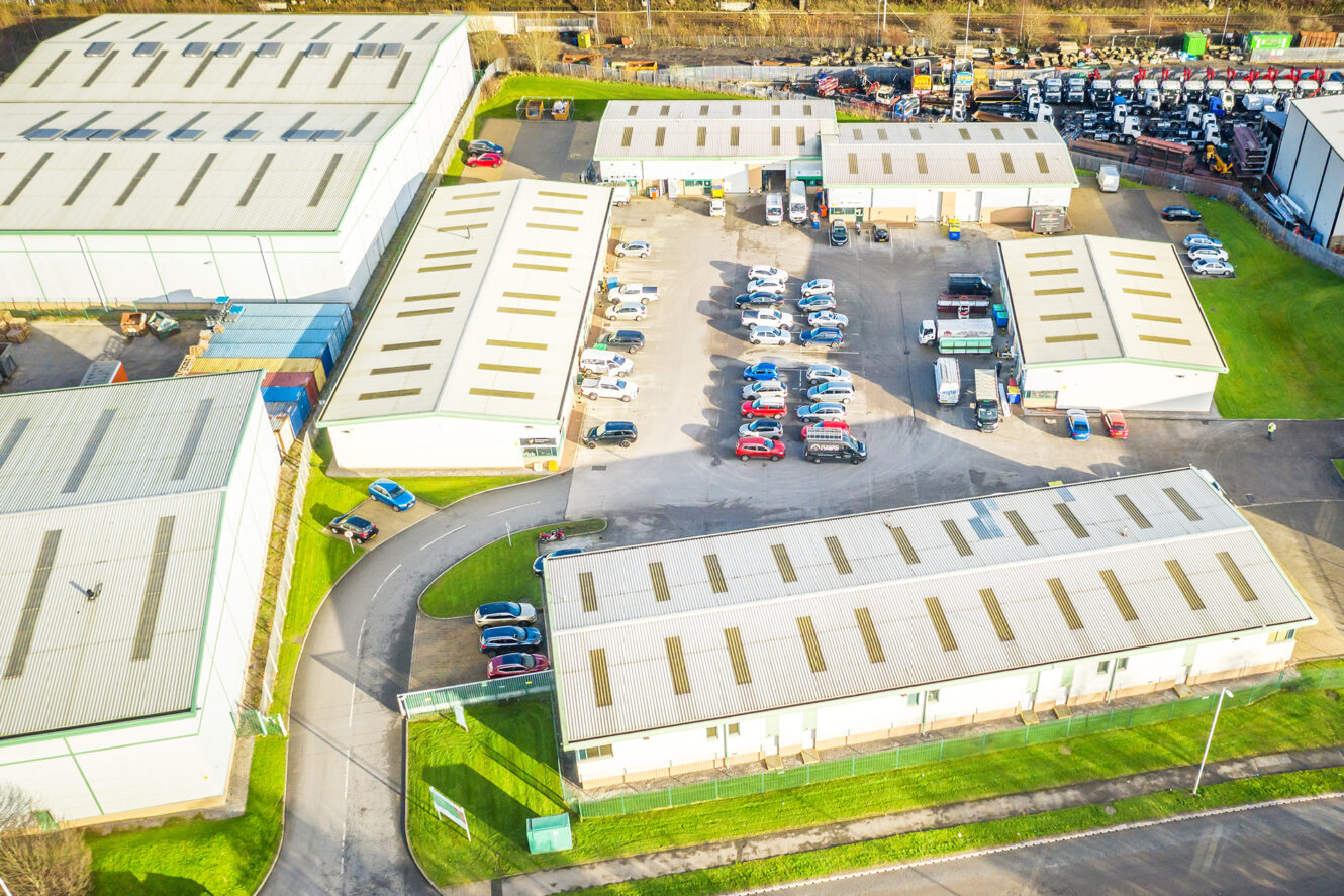 Aerial view of an industrial complex with several large warehouses, parked cars in a central lot, and trucks near a loading area.