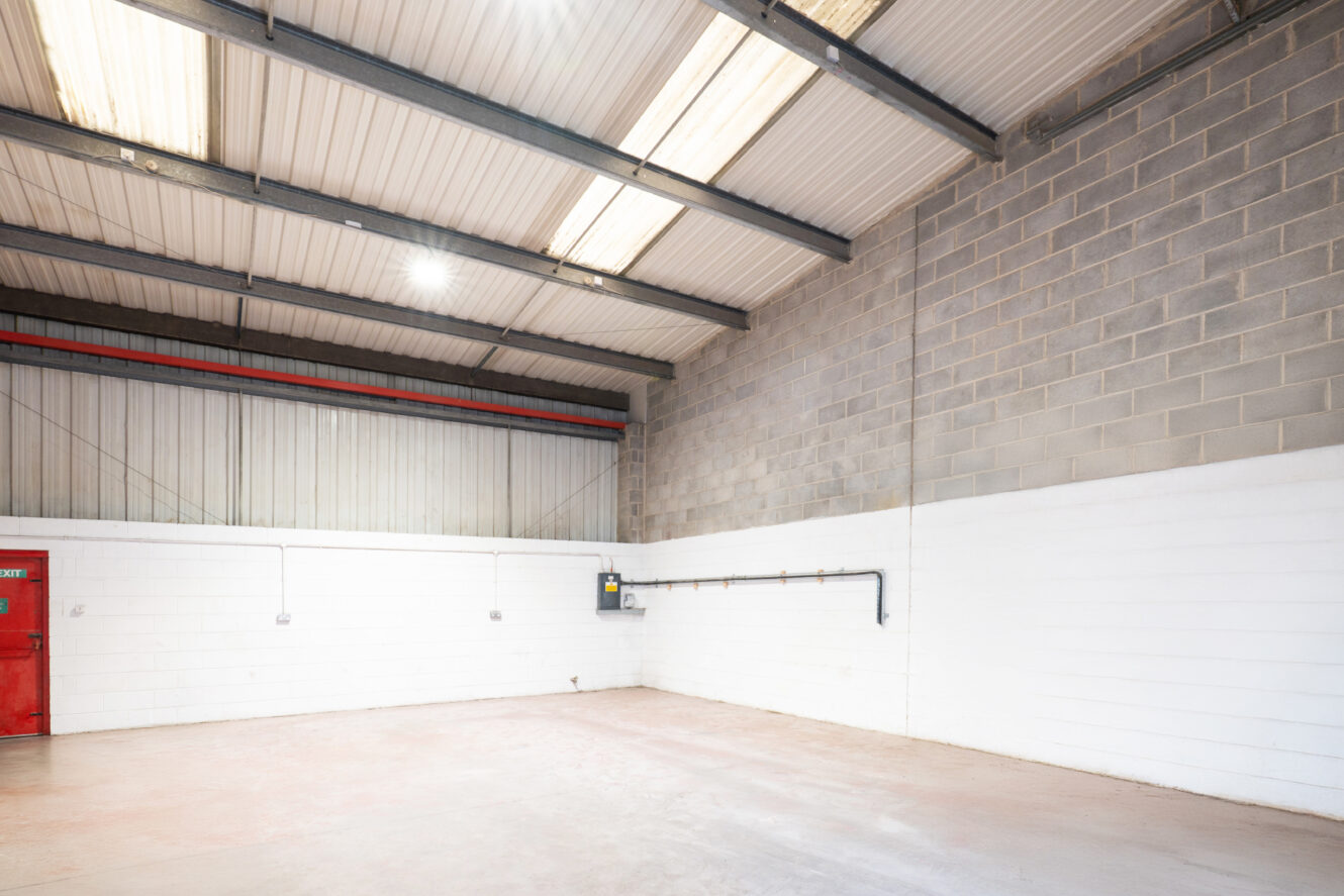 Empty industrial warehouse with a red door, white and gray brick walls, exposed pipes, and a high ceiling with metal beams and skylights.
