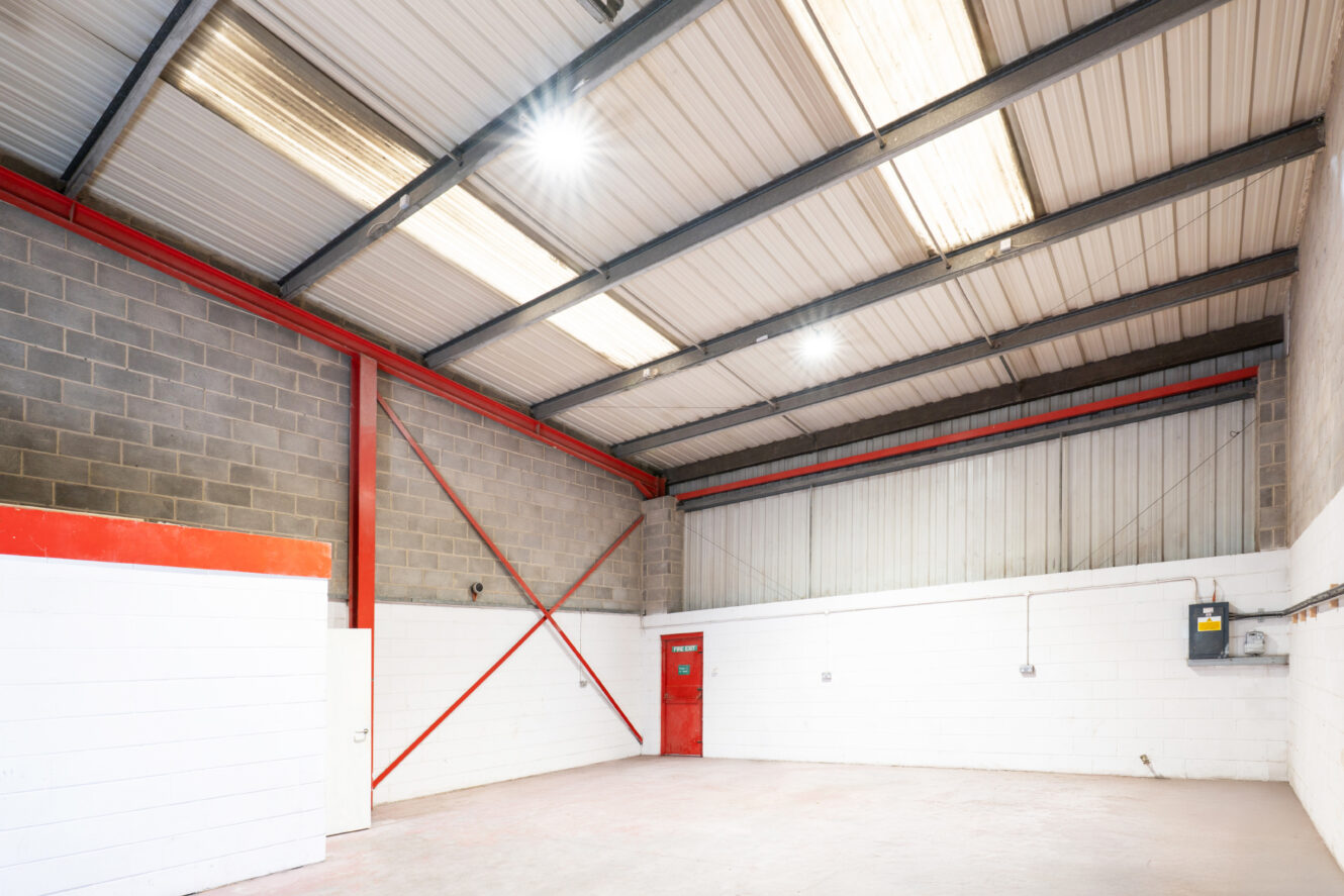 Empty warehouse interior with concrete floor, white and gray brick walls, red metal beams, a red door, overhead lights, and exposed ceiling panels.