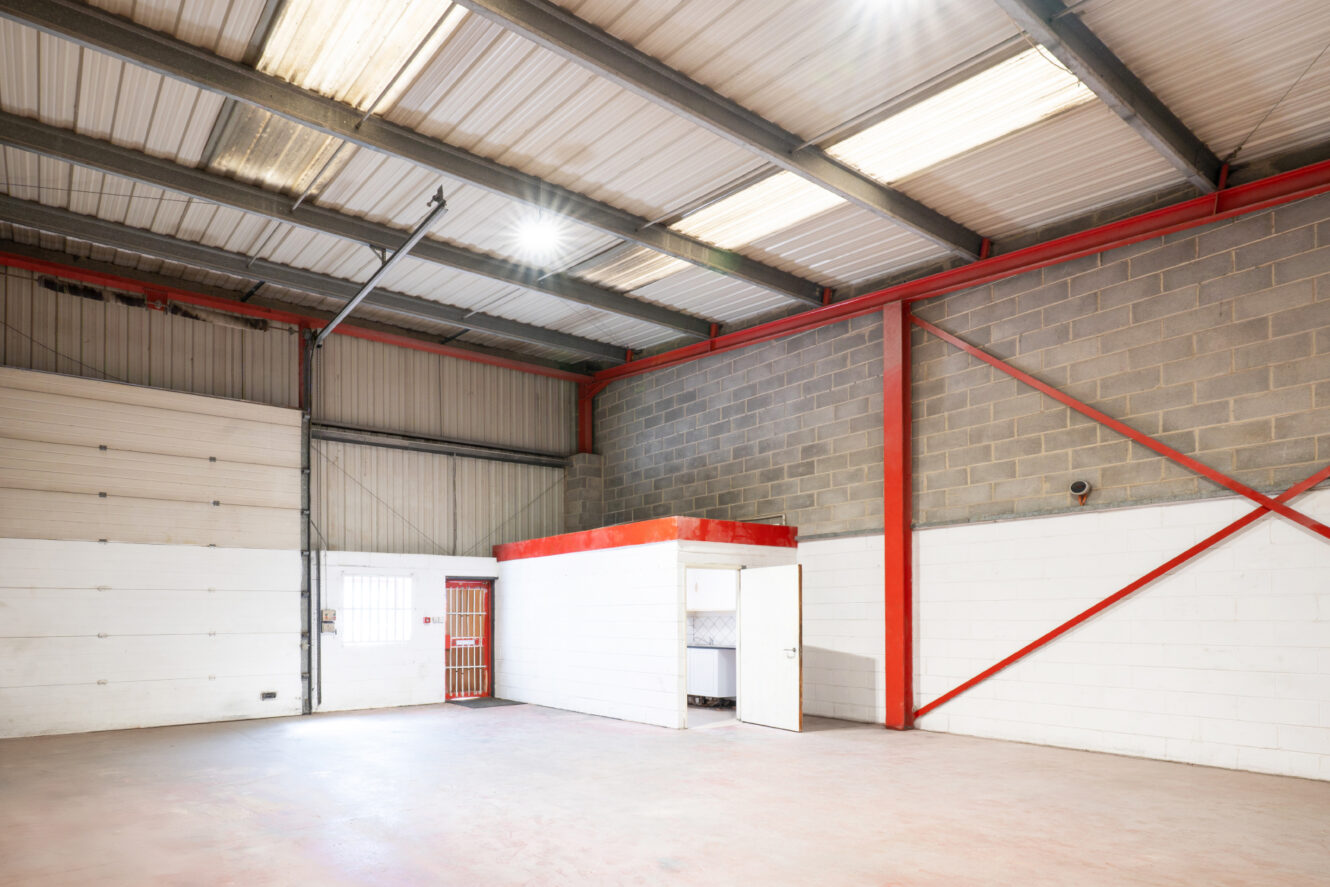 Empty industrial warehouse interior with high metal ceiling, exposed beams, concrete floor, and a small white office structure with an open door and barred window.