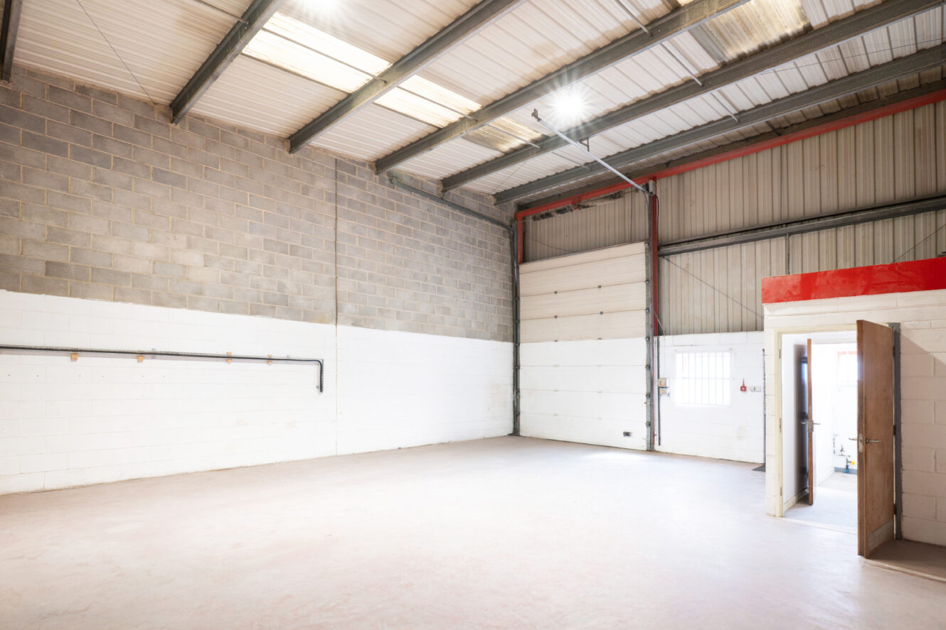 Empty industrial warehouse interior with concrete floor, high ceiling, exposed metal beams, large roller door, and small office space visible through an open door.