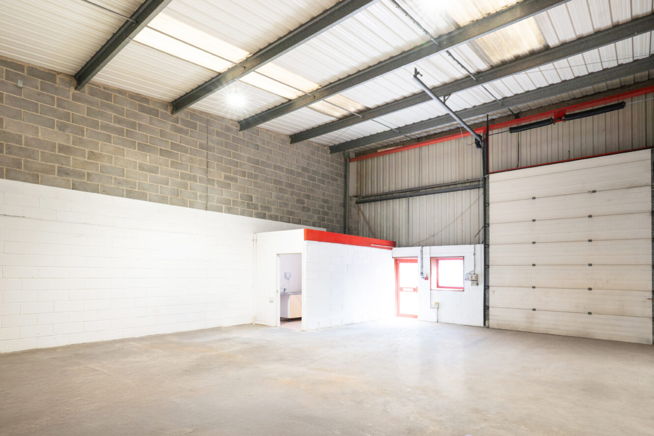 Empty industrial warehouse with high ceiling, concrete floor, brick and white walls, a small enclosed office space, and a large roller door.