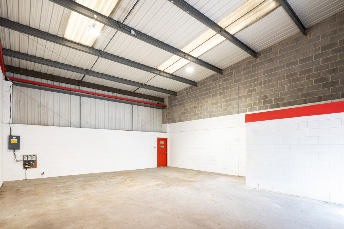 Empty industrial warehouse space with white and gray brick walls, concrete floor, metal ceiling, fluorescent lights, and a red door.