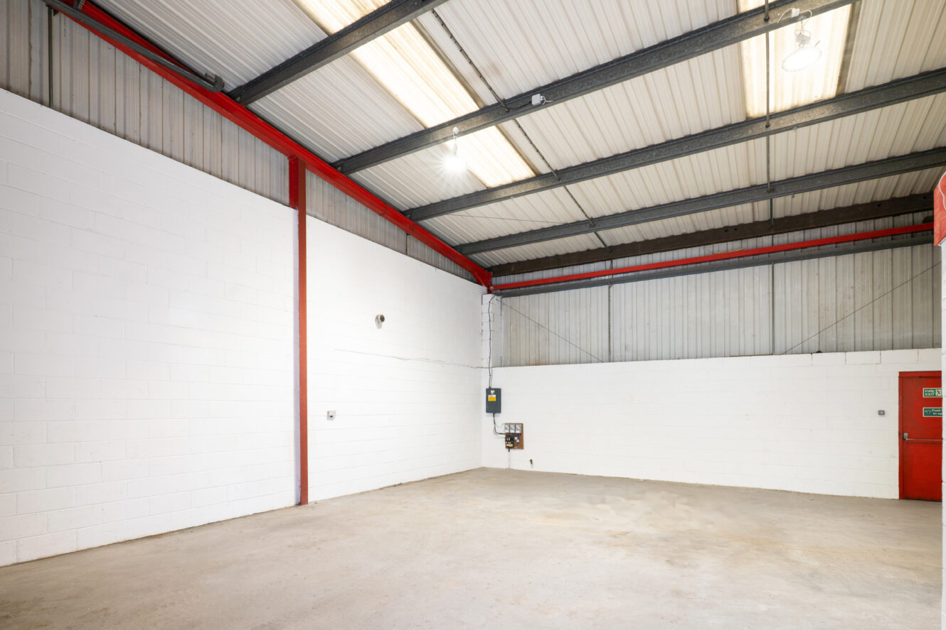 Empty industrial warehouse space with high ceilings, white brick walls, exposed metal beams, concrete floor, and electrical panels on the wall.