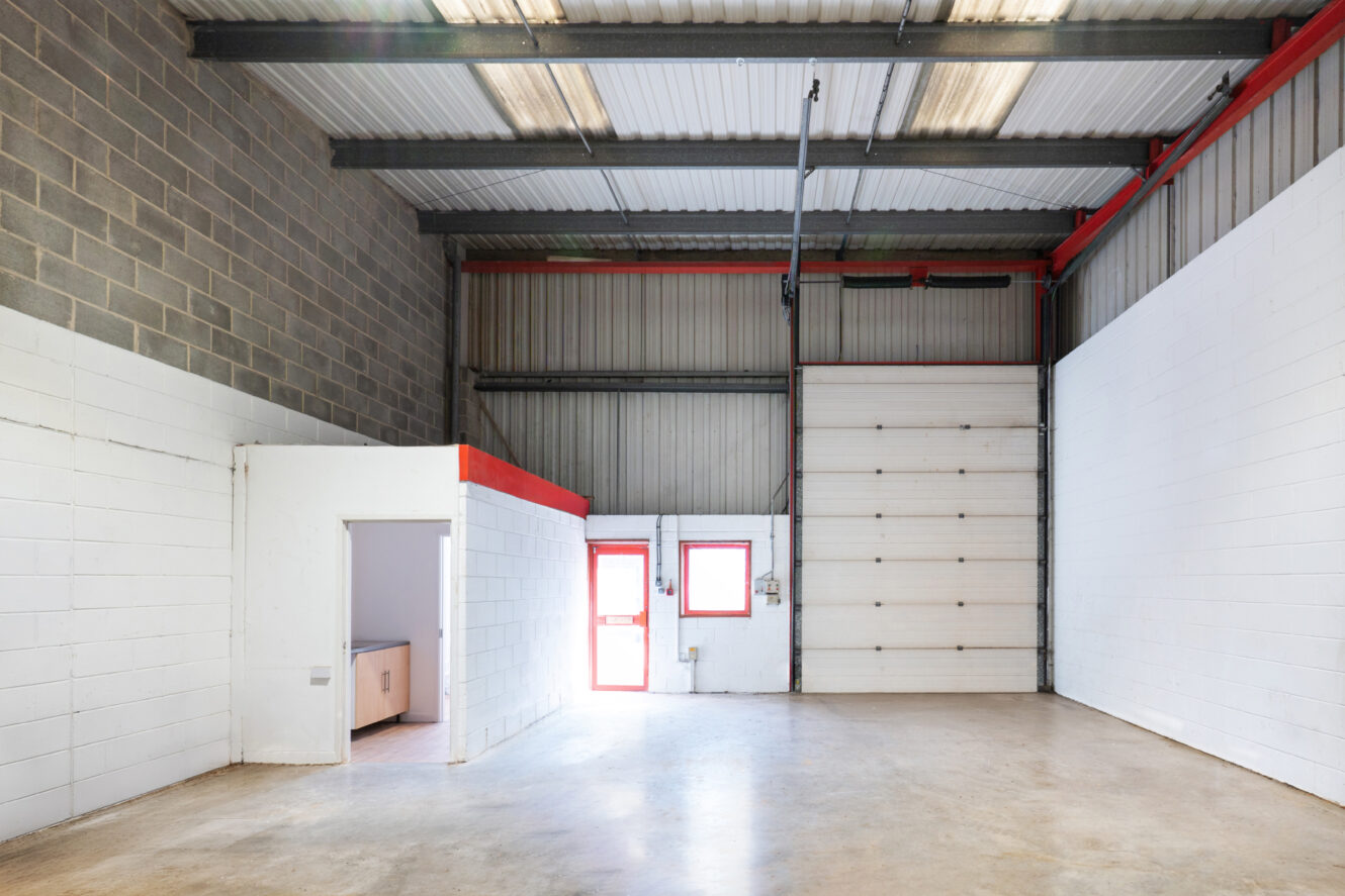 Empty industrial warehouse interior with high ceiling, white painted walls, large roller shutter door, small office area, and two red-framed windows letting in natural light.