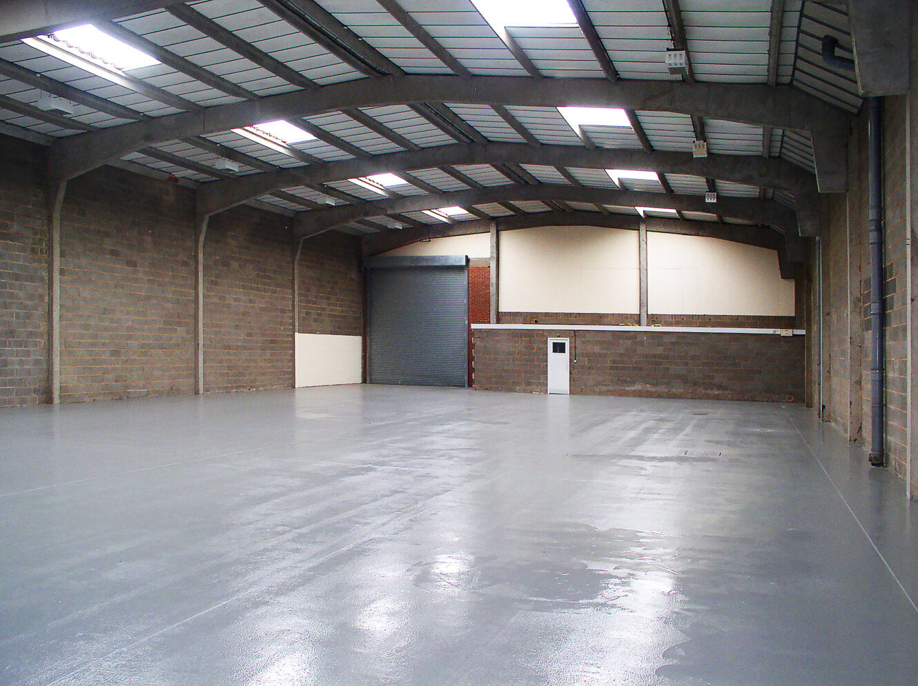 Empty warehouse interior with concrete walls, polished gray floor, exposed metal ceiling, and a large shutter door at the far end.