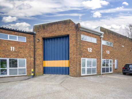 Industrial building with units numbered 11, 12, and 13. Unit 12 has a large blue and orange roller shutter door. A black car is parked next to unit 13.