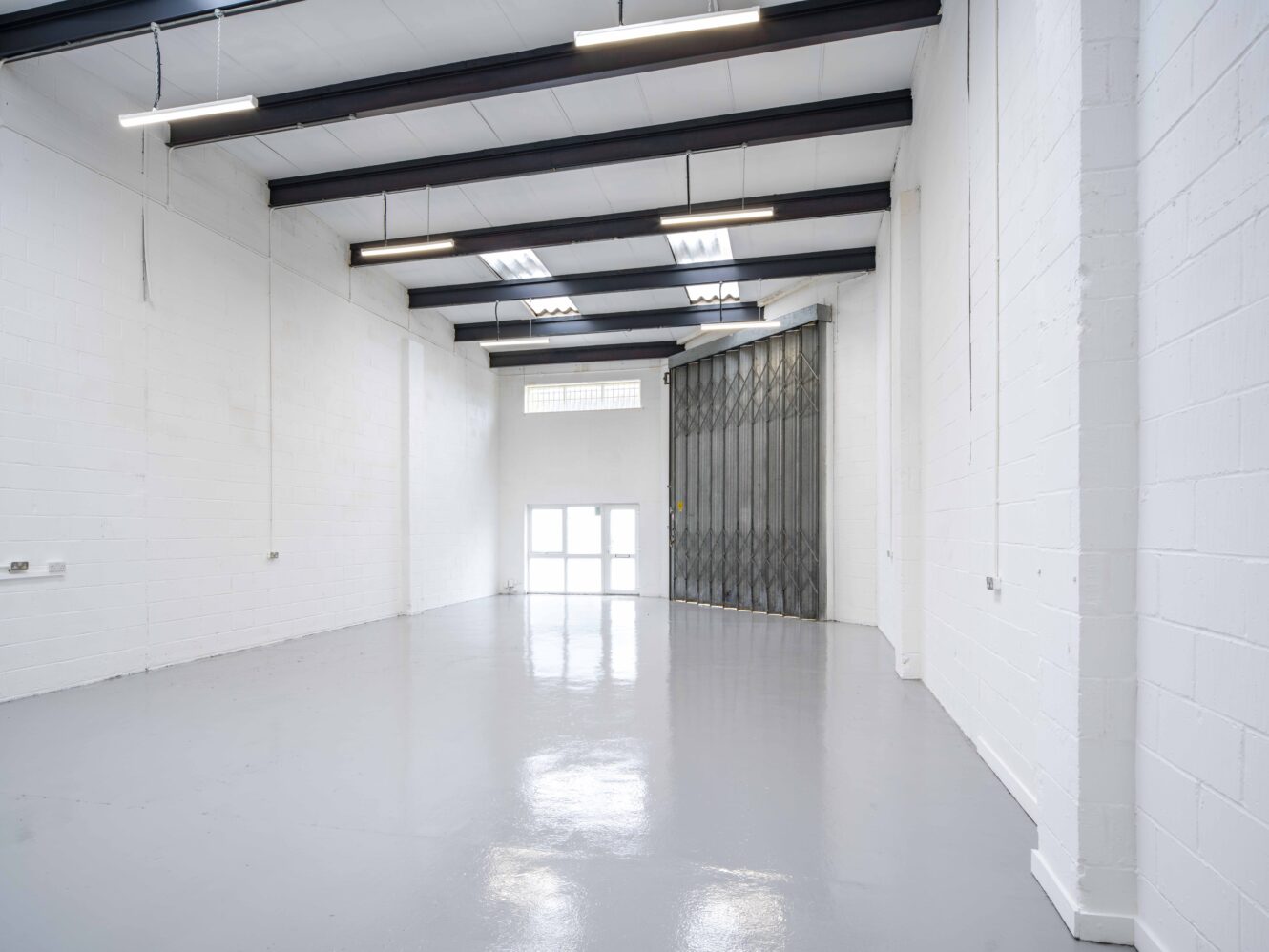 Empty industrial warehouse unit with white painted walls, grey polished floor, high ceiling with exposed beams, fluorescent lights, and a large metal folding door.