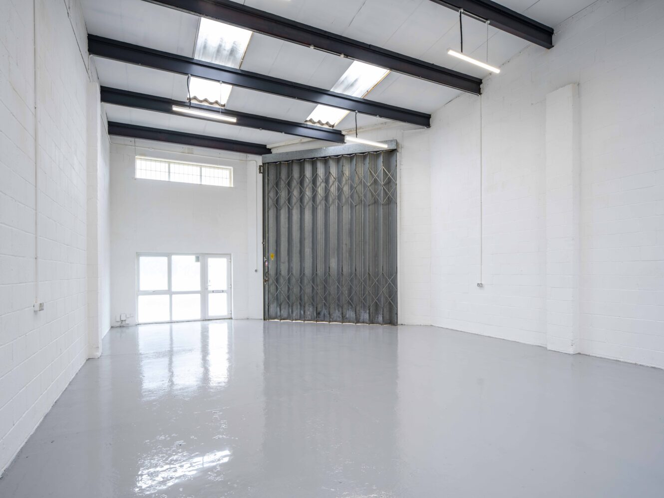 Empty industrial warehouse with white painted walls, polished concrete floor, metal ceiling beams, large folding metal door, and a window letting in natural light.