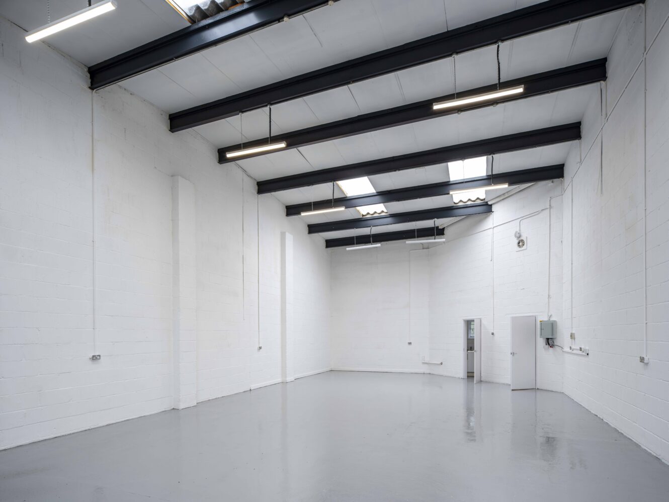 Empty industrial warehouse with white painted brick walls, gray concrete floor, exposed steel beams, and fluorescent lighting. A door and electrical panels are visible on the right wall.