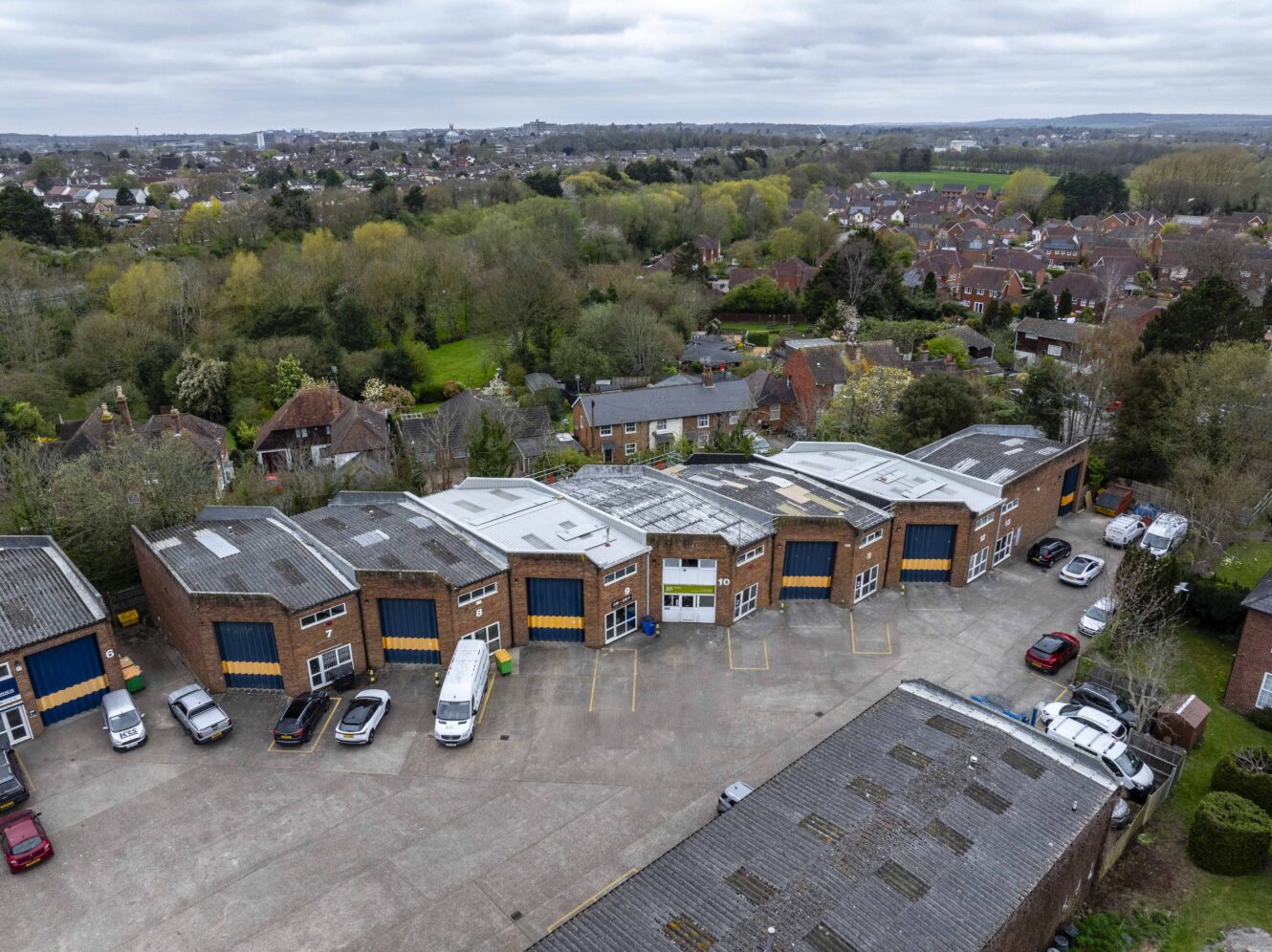 Aerial view of a row of industrial units with vehicles parked outside, surrounded by trees and residential houses in the background under a cloudy sky.