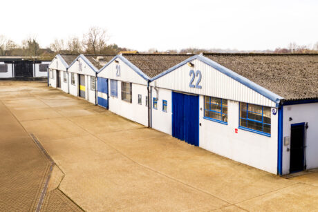 Row of industrial warehouse units with white and blue exteriors, large numbers on each unit, and a concrete yard in front.