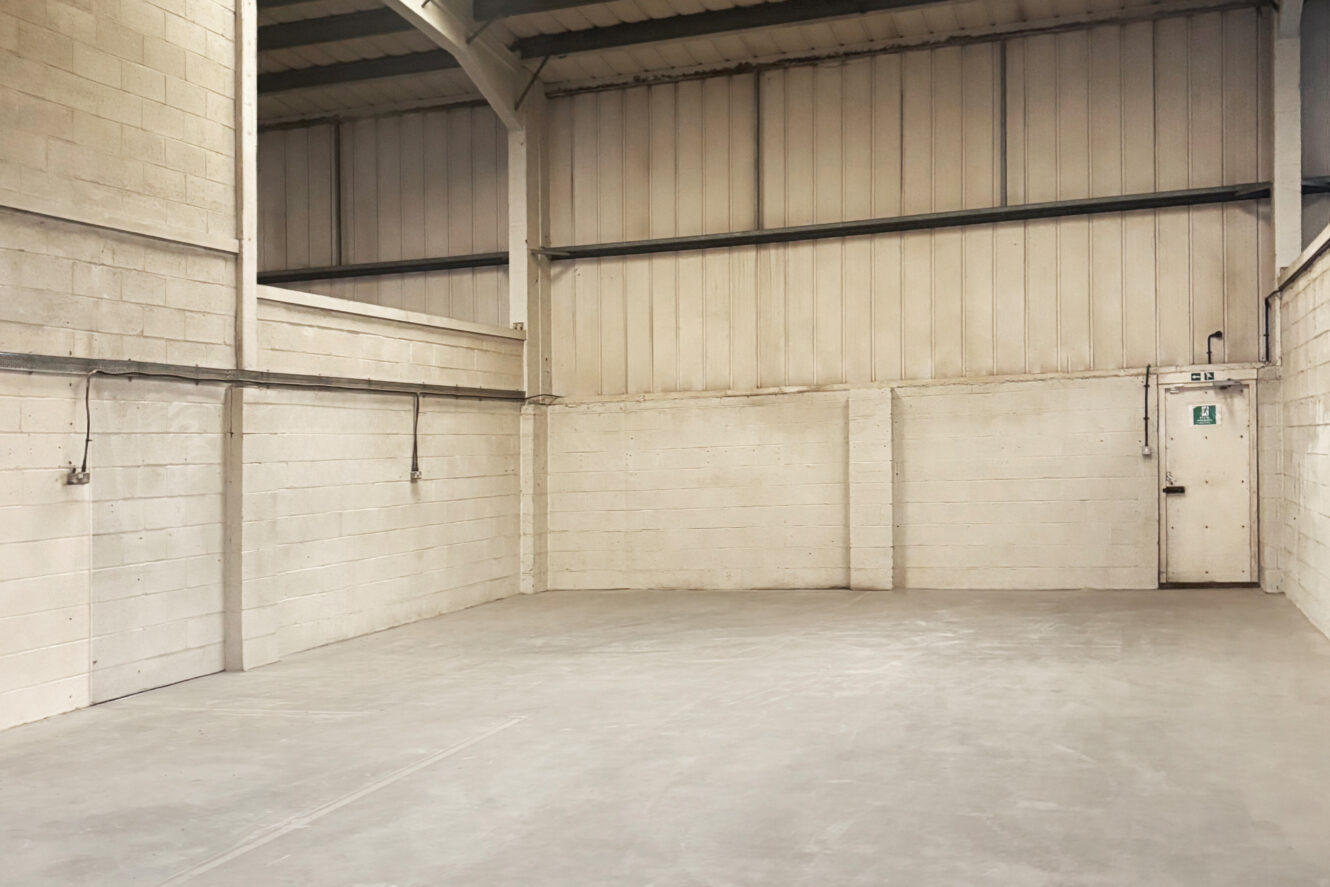 Empty industrial warehouse with concrete floor, white brick walls, high ceiling, and a closed metal door with an emergency exit sign.
