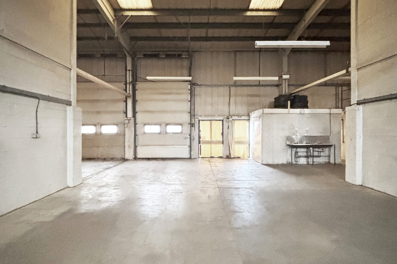 Empty industrial warehouse interior with concrete floor, white walls, high ceiling, fluorescent lights, small enclosed room, sinks, and large garage door illuminated by sunlight.