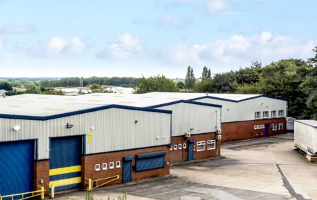 A row of industrial warehouse buildings with blue roller doors and brick exteriors, surrounded by paved areas and trees in the background.