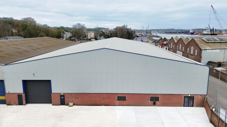 Large industrial warehouse with a gray metal roof, red brick lower walls, and a black roller door, situated near a road and waterway with cranes in the background.