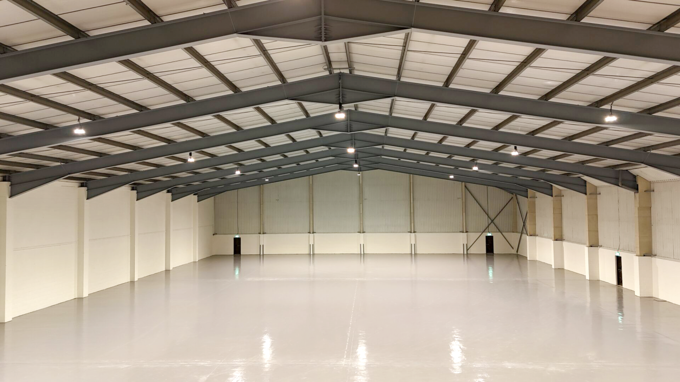 Interior of an empty warehouse with a polished concrete floor, high ceiling with exposed beams, and evenly spaced overhead lights.