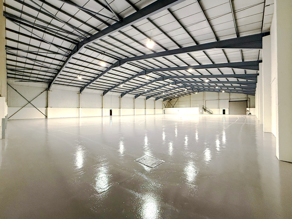 Large, empty warehouse with high ceilings, exposed steel beams, white walls, and a glossy, reflective floor under bright overhead lights.