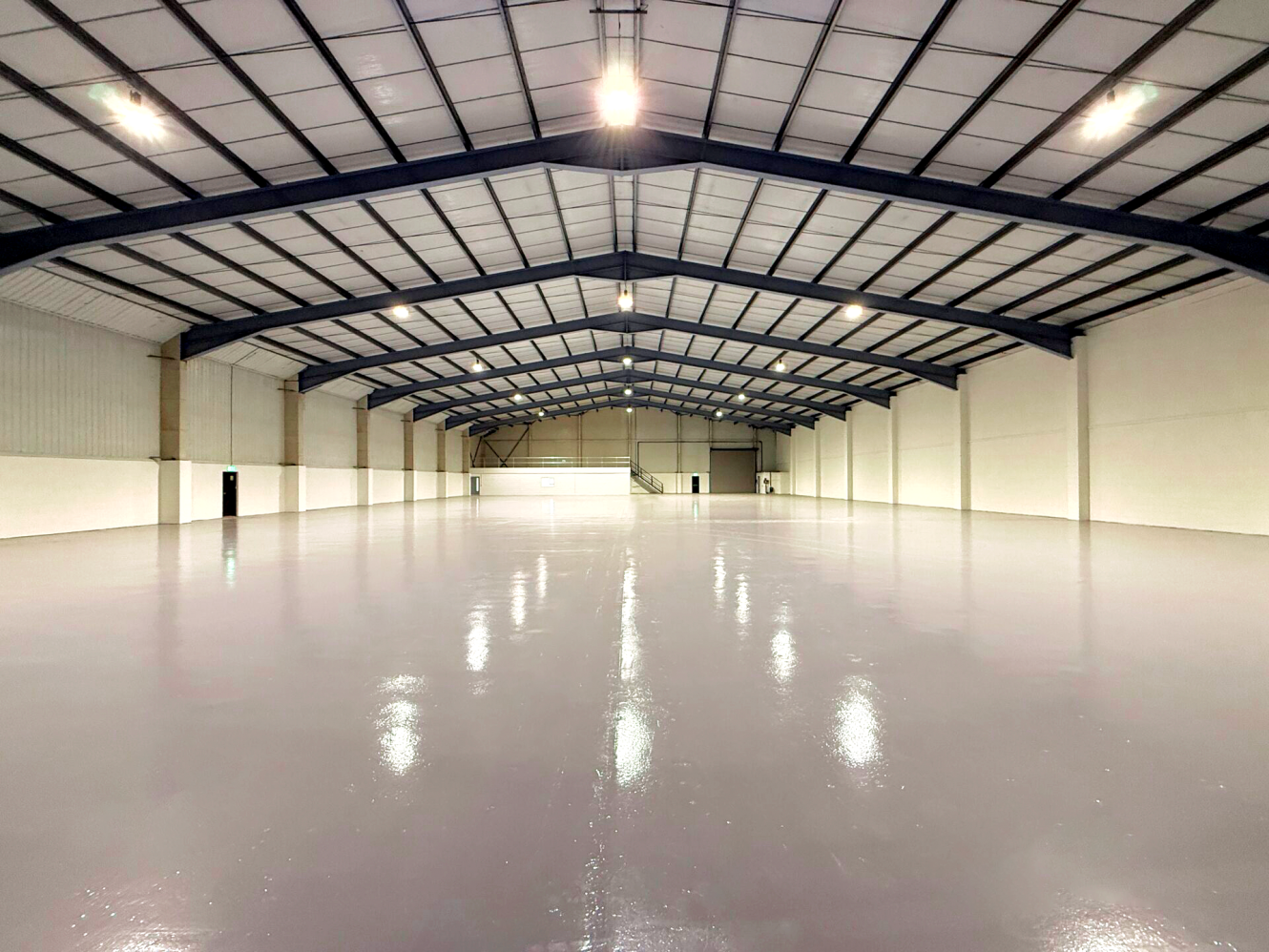 Large, empty warehouse with a shiny polished floor, white walls, and a high ceiling supported by steel beams.