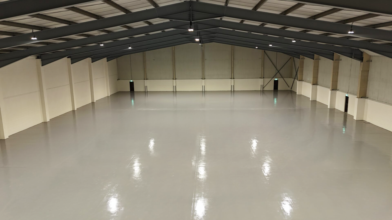Spacious, empty warehouse with high ceilings, white walls, and polished concrete floor, illuminated by overhead lights. Multiple exit doors visible along the side and end walls.
