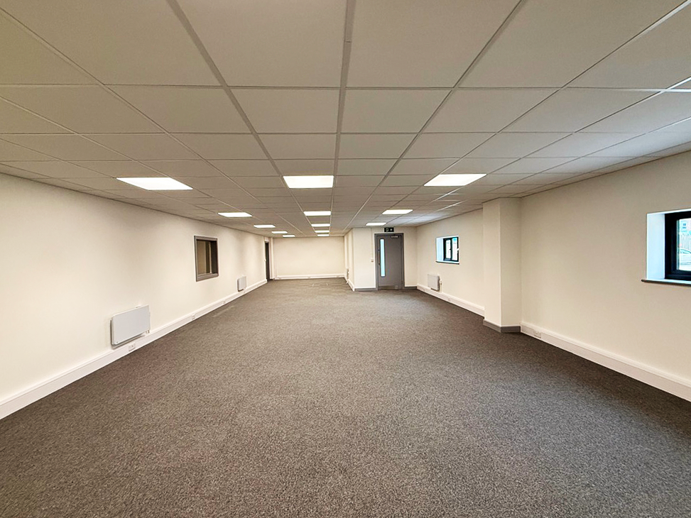 Empty office space with white walls, grey carpet, several windows, ceiling lights, and a closed door at the far end.