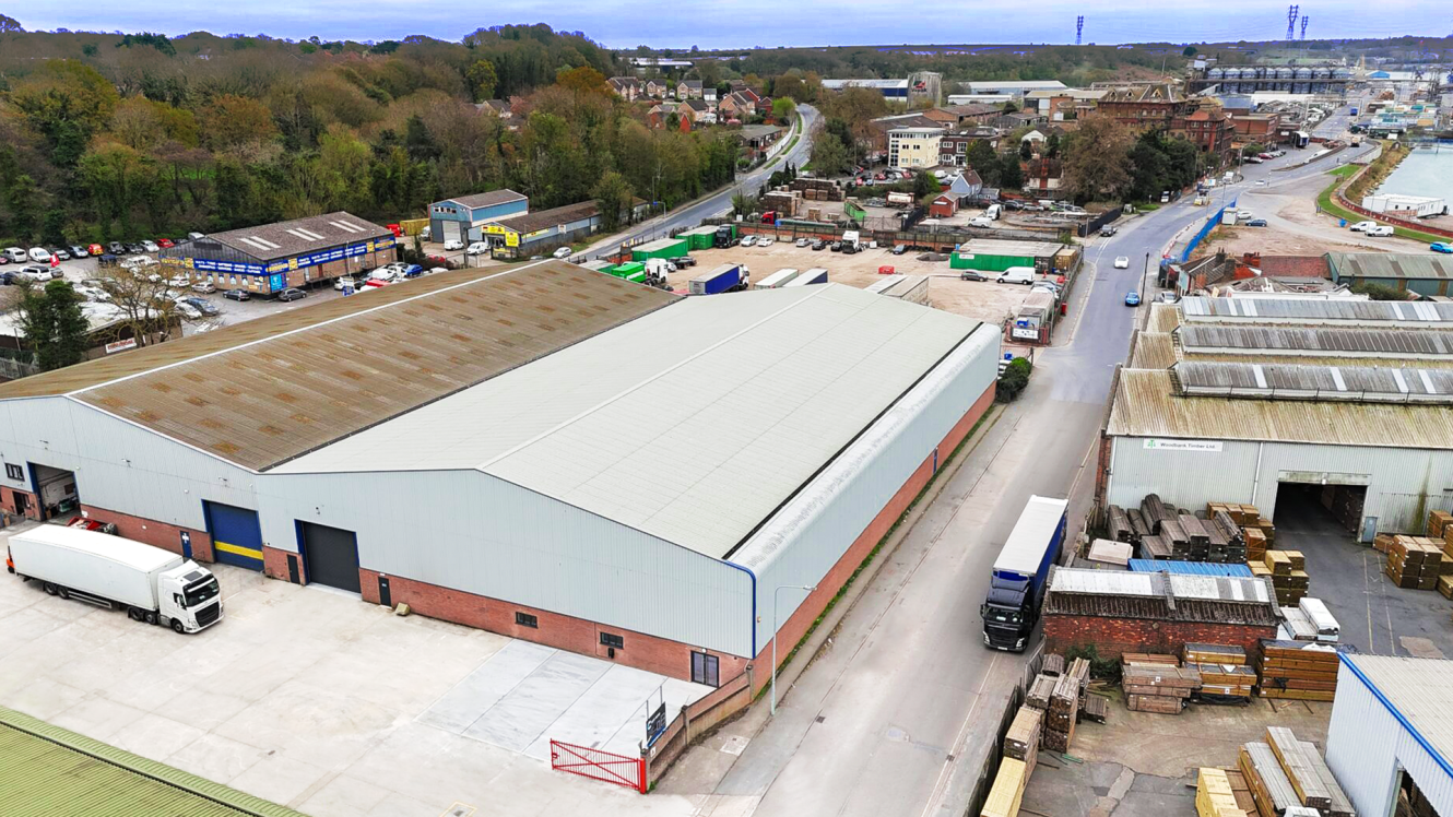 Aerial view of an industrial warehouse complex with trucks parked outside, surrounded by roads, trees, and additional buildings.
