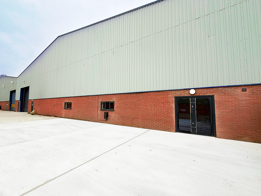 Exterior view of a large industrial warehouse with a brick lower facade, metal upper walls, and glass double doors, set on a wide concrete lot.