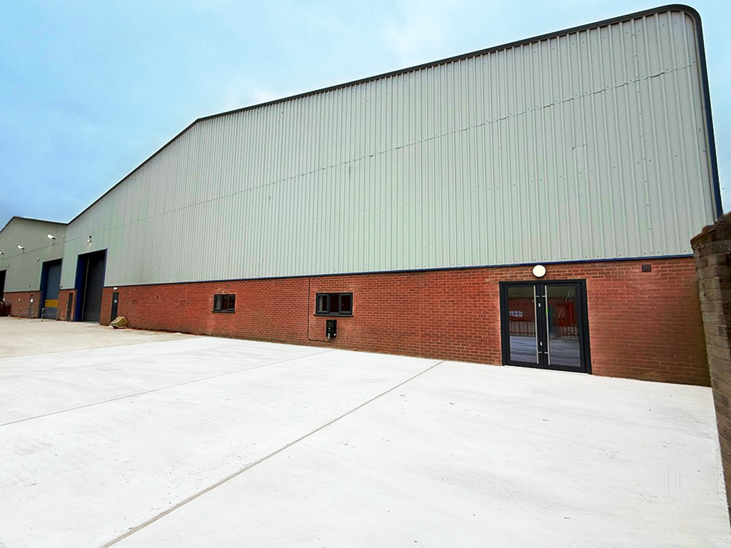 A large industrial warehouse with a metal upper facade and brick lower walls, featuring several doors and windows, with a wide concrete lot in front.