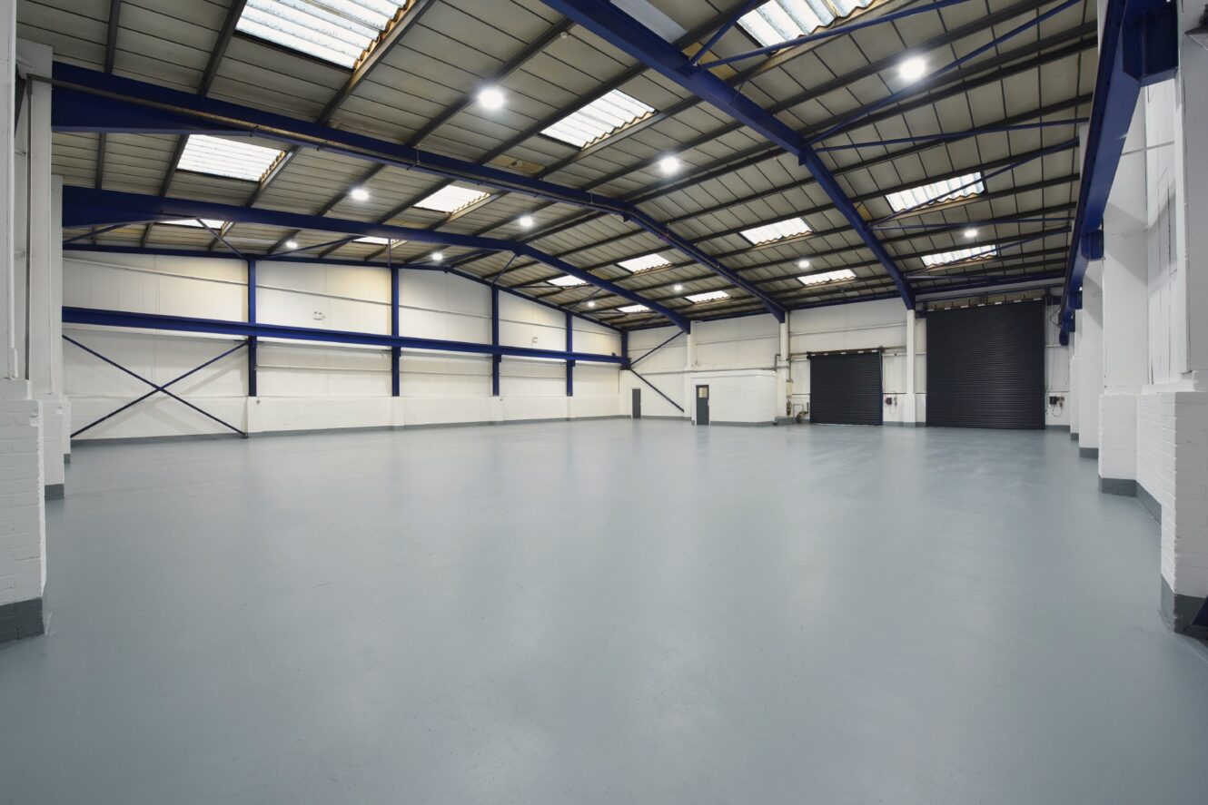Spacious, empty industrial warehouse with high ceilings, white walls, blue beams, and skylights. Large roller door visible on one side.