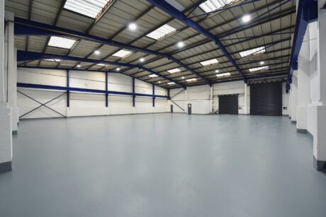 Spacious, empty industrial warehouse with high ceilings, white walls, blue beams, and skylights. Large roller door visible on one side.