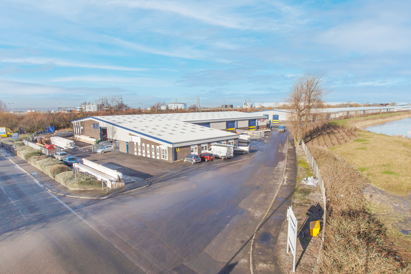 A wide view of an industrial warehouse complex with trucks parked outside, surrounded by paved roads, fencing, and open grassy areas under a blue sky.