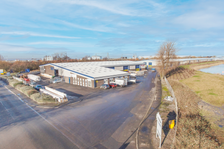 A wide view of an industrial warehouse complex with trucks parked outside, surrounded by paved roads, fencing, and open grassy areas under a blue sky.