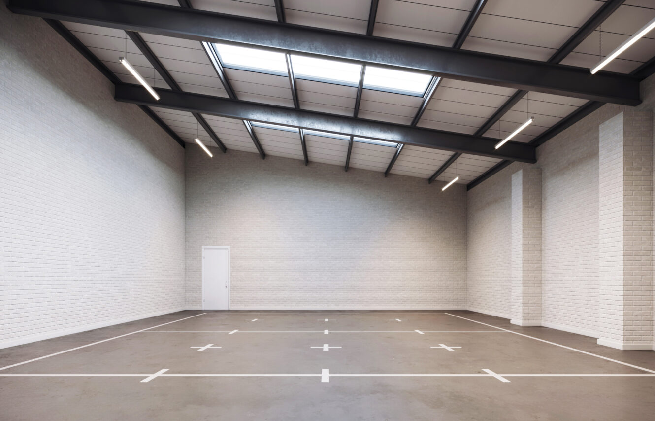 Empty indoor warehouse with white brick walls, exposed beams, skylights, and marked sections on the concrete floor; a single door is visible on one wall.