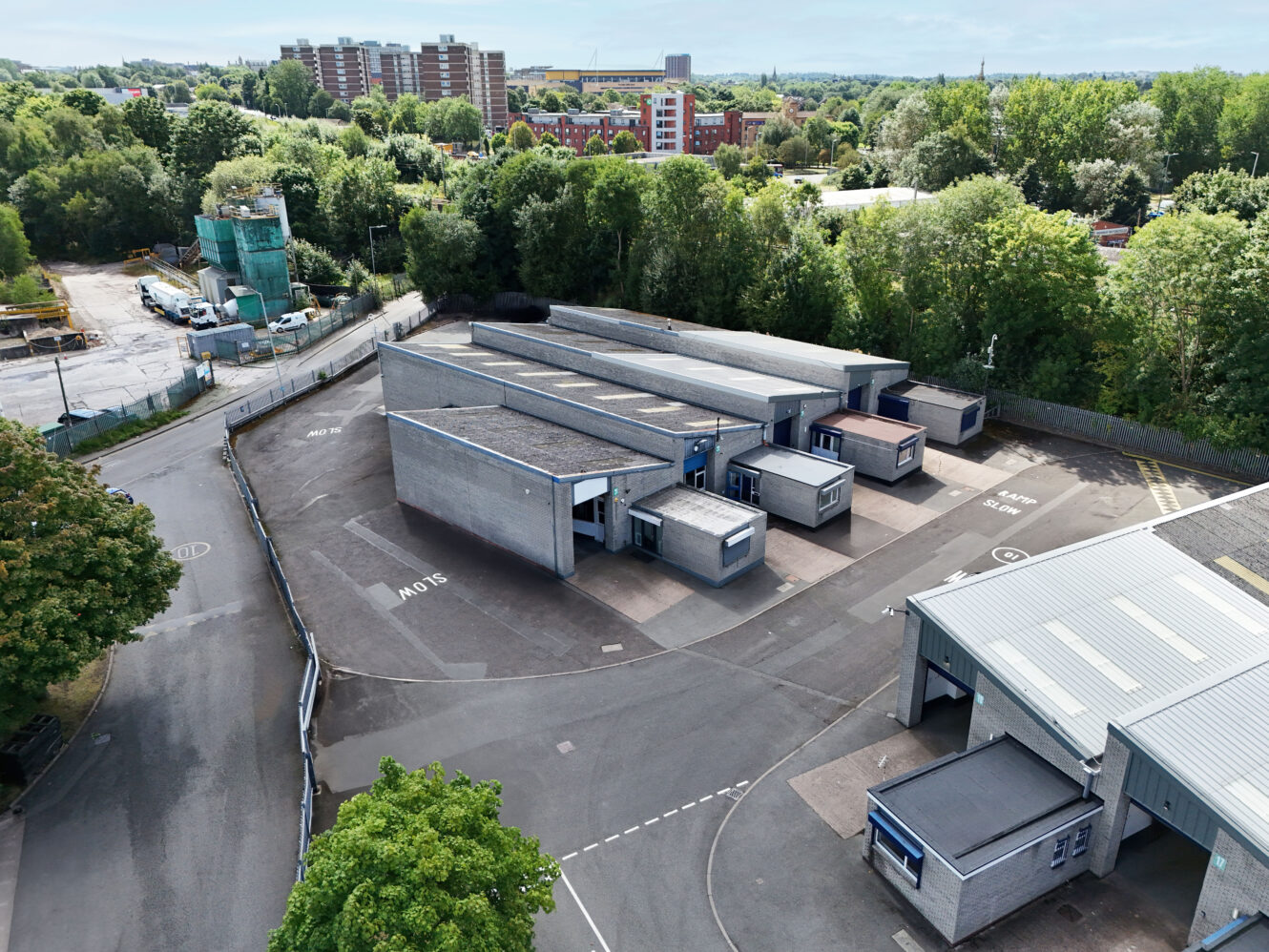 Aerial view of an industrial complex with several rectangular gray buildings surrounded by trees, parking areas, and adjacent roads. Residential buildings are visible in the background.