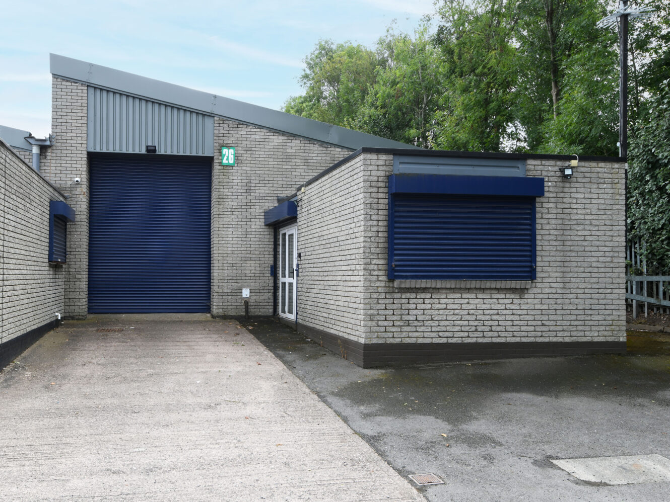 A light gray brick industrial building with blue roller shutters, a driveway, a closed door, and trees in the background.