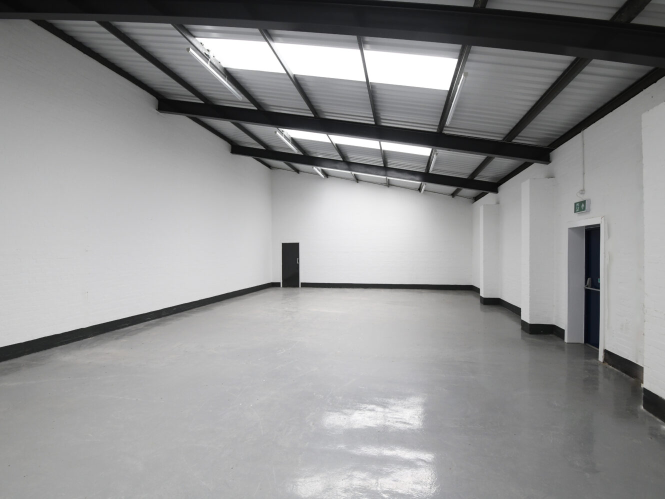 Empty industrial warehouse space with white walls, a smooth gray floor, black metal beams, overhead skylights, a small door at the back, and an open doorway on the right.