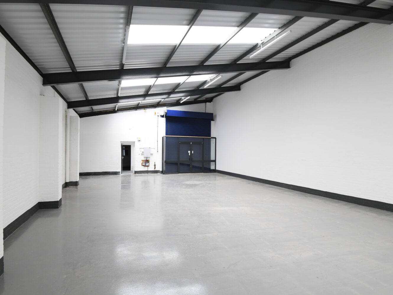 Empty industrial warehouse with white painted walls, grey polished floor, metal ceiling beams, and double glass doors at the far end.