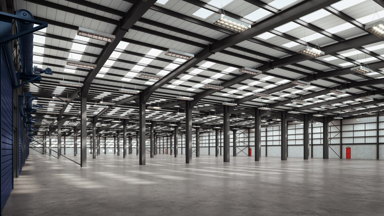 Interior of a large empty warehouse with metal beams, concrete floor, skylights, and bright overhead lighting. Several red doors line the far wall.