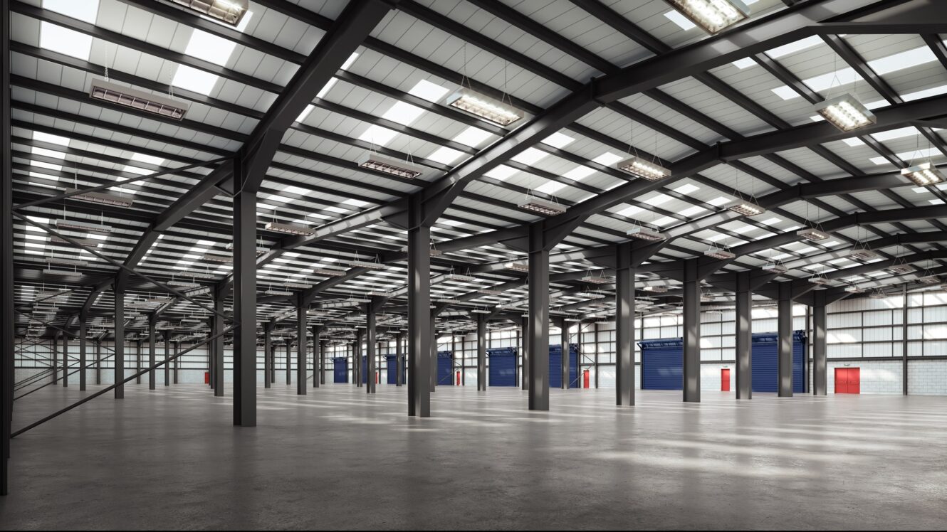 Interior of a large, empty warehouse with concrete floors, steel beams, high ceilings, and several blue and red rolling shutter doors along the walls.