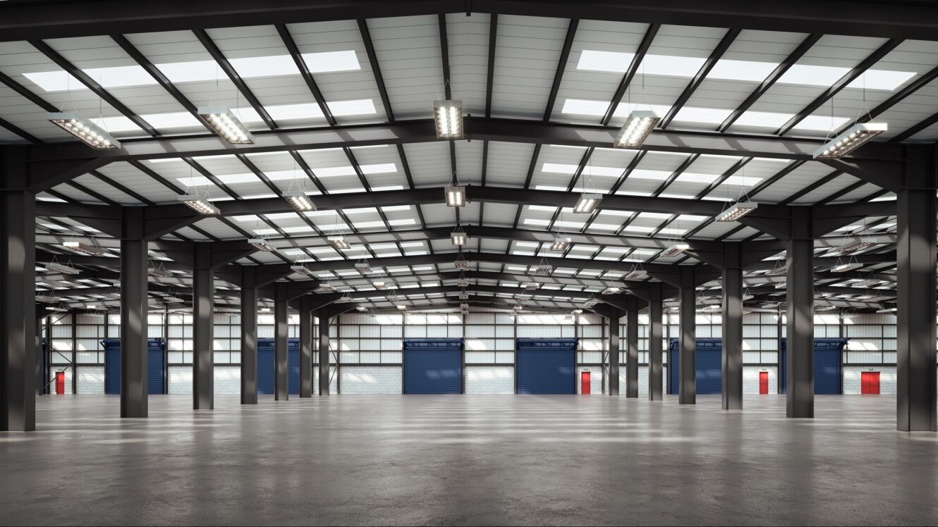 Interior of an empty, modern warehouse with high ceilings, metal beams, large skylights, and several closed blue roller doors along the back wall.