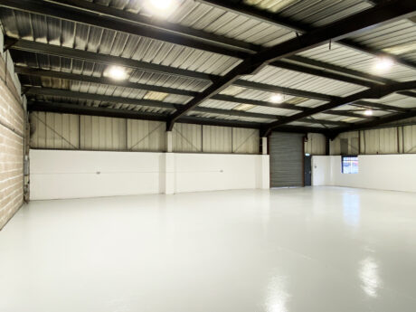 Empty industrial warehouse with clean white floors, metal walls, exposed beams, overhead lights, a large roller door, and a small window.