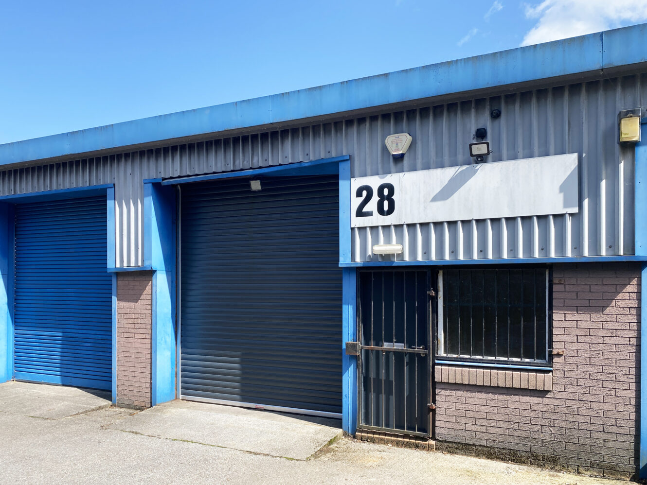 Industrial unit with blue trim, two roller doors, one open, one closed, number 28 sign above, and a barred window on a brick and metal exterior.