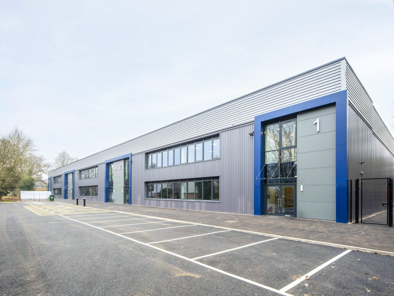 A modern industrial or office building with large windows, metal siding, and an empty parking lot in front. The building is labeled with the number 1.
