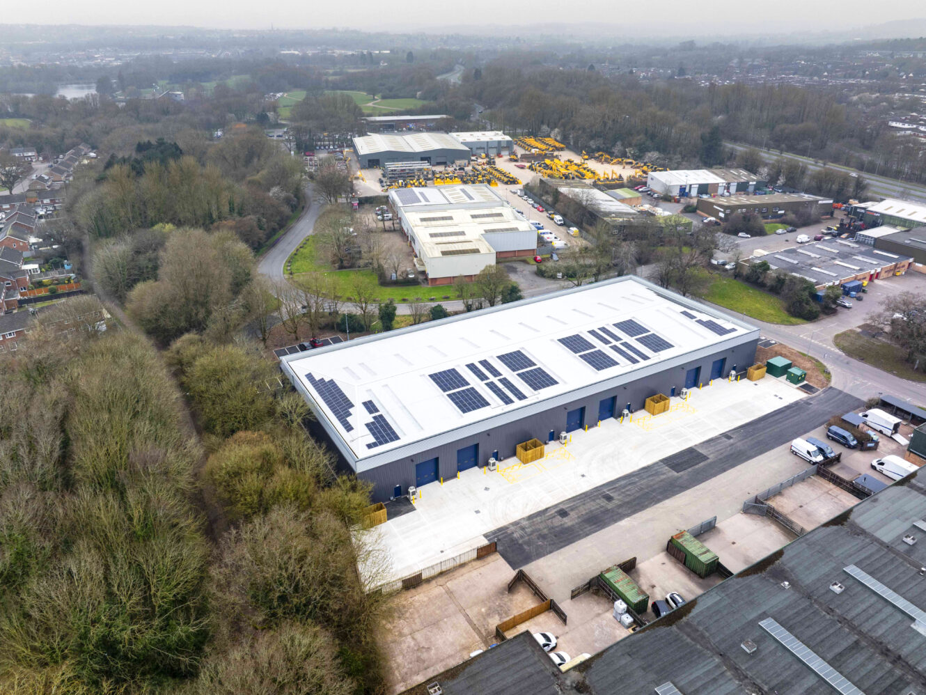 Aerial view of a large industrial building with solar panels on the roof, surrounded by trees and other industrial facilities.