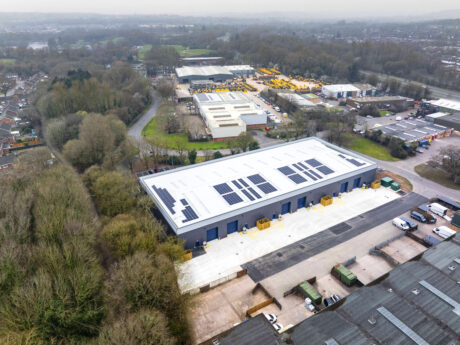 Aerial view of a large industrial building with solar panels on the roof, surrounded by trees and other industrial facilities.