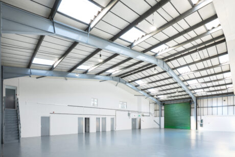 Spacious, empty warehouse with high ceilings, skylights, white walls, grey doors, a staircase, and a large green industrial shutter door.