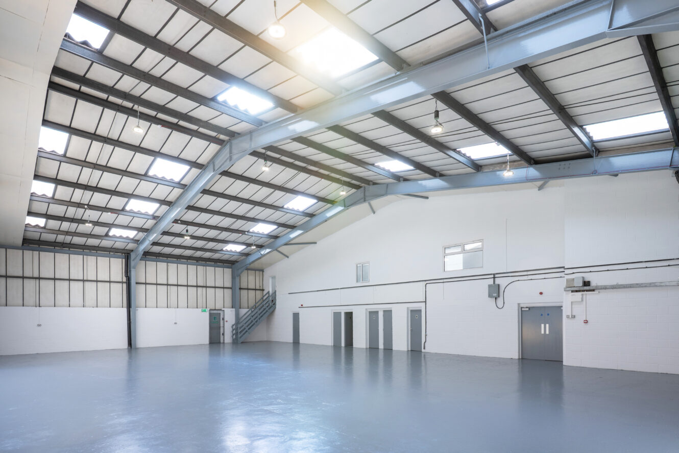 Large, empty industrial warehouse with high ceilings, metal beams, polished concrete floor, and multiple doors along the white walls. Natural light enters through skylights.